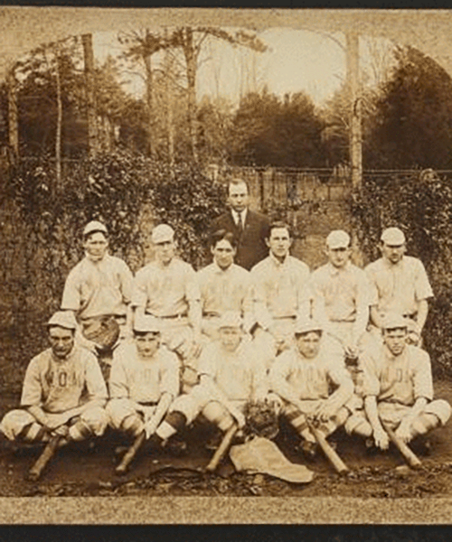 Vintage sepia photograph of a baseball team with nine players and one coach, all in traditional uniforms and caps, posed with bats and gloves in a serene outdoor setting with trees and a bridge in the background.
