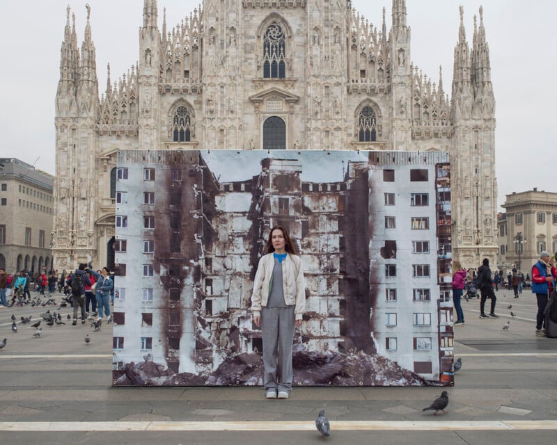 A person stands in front of a large photo showing a collapsed building, set up in a public plaza. The stunning Gothic architecture of the Milan Cathedral looms in the background. Pigeons and other people are scattered across the square.