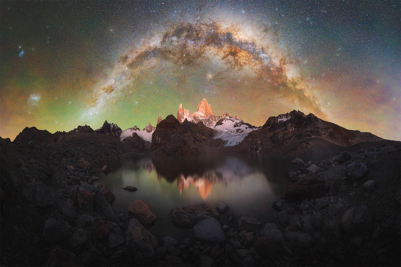 A breathtaking night sky over a mountainous landscape with a vibrant Milky Way arc stretching across the sky. Snow-capped peaks are reflected in a calm lake in the foreground, enhancing the serene and majestic atmosphere.