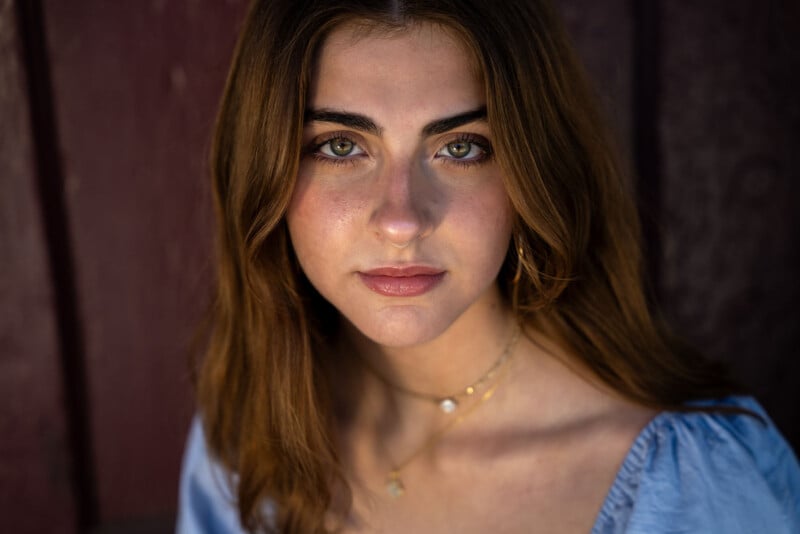 A young woman with long, light brown hair and green eyes looks directly at the camera. She is wearing a light blue blouse and a gold necklace with small pendants. The background is dark and slightly blurred, highlighting her face.