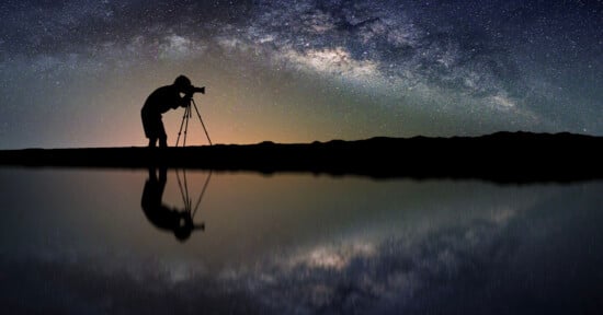 Silhouette of a person using a camera on a tripod against a starry night sky backdrop. The nightscape includes a vivid Milky Way galaxy. The scene is reflected perfectly on a calm body of water below, creating a symmetrical and serene composition.