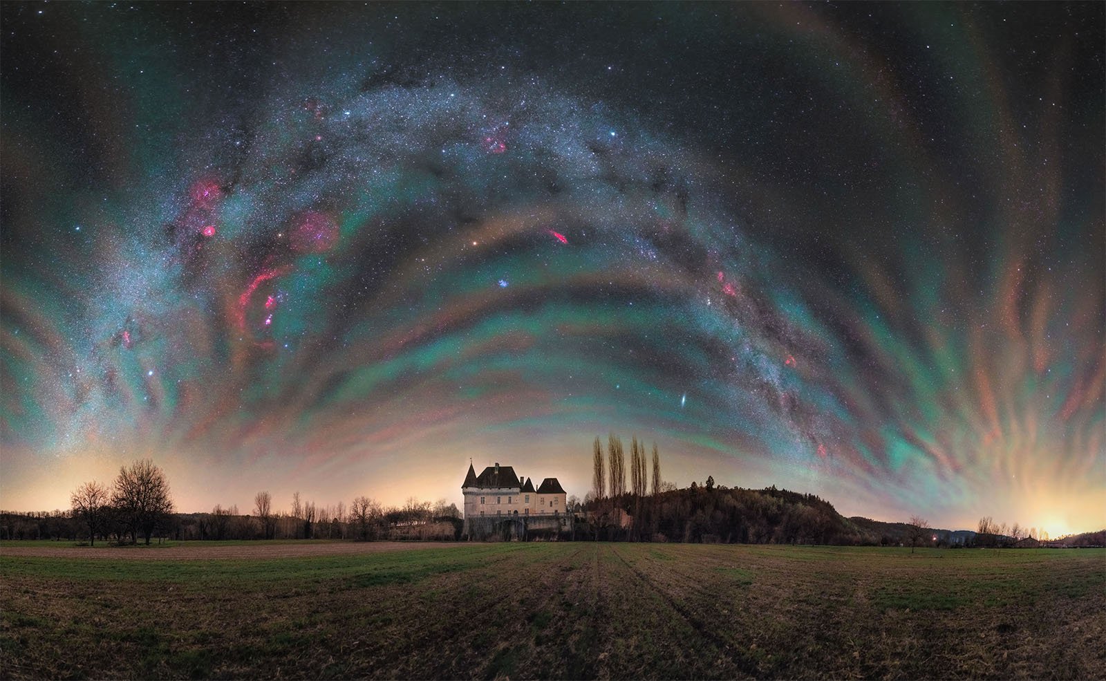 A panoramic night sky showcases stunning auroras and a colorful Milky Way arching over a landscape with an old mansion. The field in the foreground and distant trees create a serene rural setting under the celestial display.