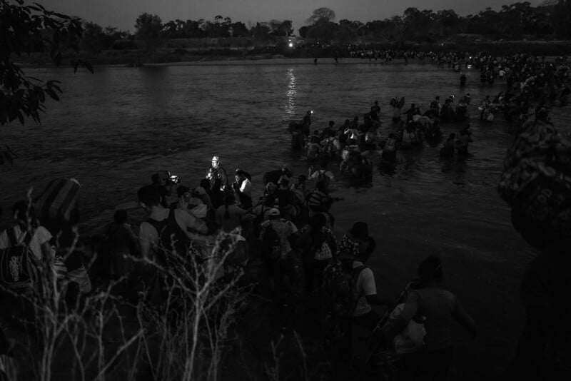 A nighttime scene of a crowded riverbank where people, illuminated by handheld lights, wade and gather in the water, with more spectators lining the shore.