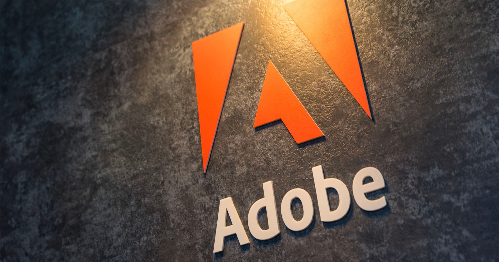 Close-up of the adobe logo with a stylized "a" in orange above the word "adobe" in white, affixed to a textured dark gray wall.