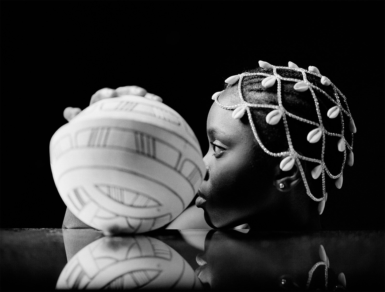 A black and white photo of a child with decorative beads in her hair touching noses with a striped, textured ball, both reflected on a glossy surface.