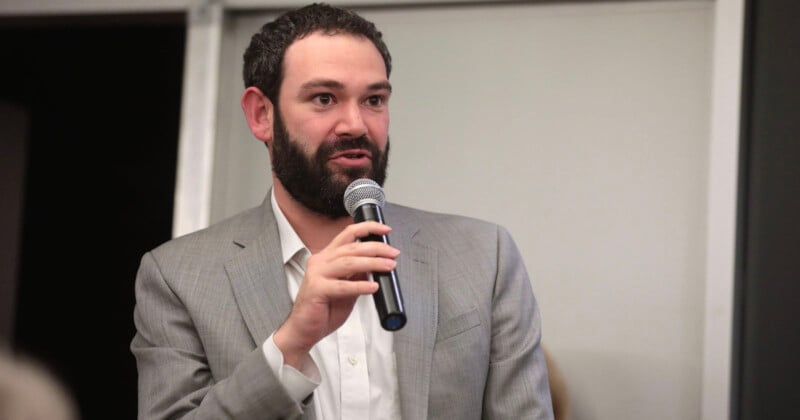 A man with short, dark curly hair and a beard is standing and speaking into a microphone. He is wearing a light grey blazer over a white dress shirt. The background is blurred, with a visible doorframe.