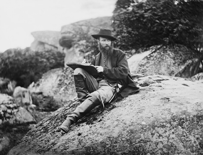 A bearded man wearing a wide-brimmed hat and 19th-century attire is sitting on a large rock in an outdoor setting. He is holding a sketchpad and appears to be drawing or writing. The background features large rocks and foliage.
