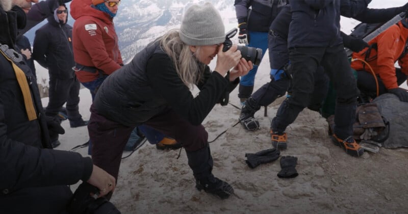 A group of people dressed in winter gear are on a snowy mountain. One person in the foreground is crouched down, taking a photograph with a camera. Some other individuals appear to be preparing for an activity or adjusting equipment. Snow-covered mountains are visible in the background.
