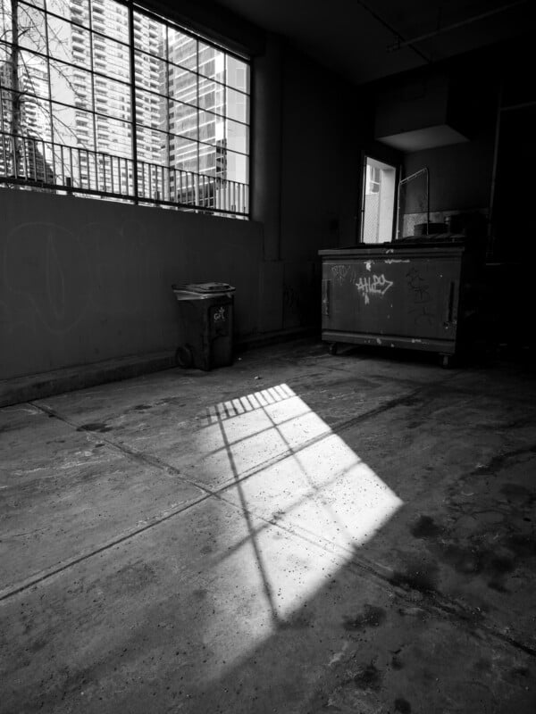 Black and white photo of a dimly lit room with a window casting shadow patterns on the floor. a trash bin and a large dumpster, both tagged with graffiti, are visible.