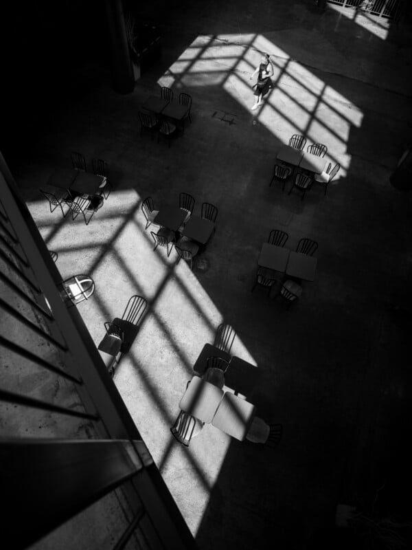A person sits alone at a table in a dimly lit cafeteria with rays of light casting shadows through large windows, highlighting a pattern of empty chairs and tables around them.