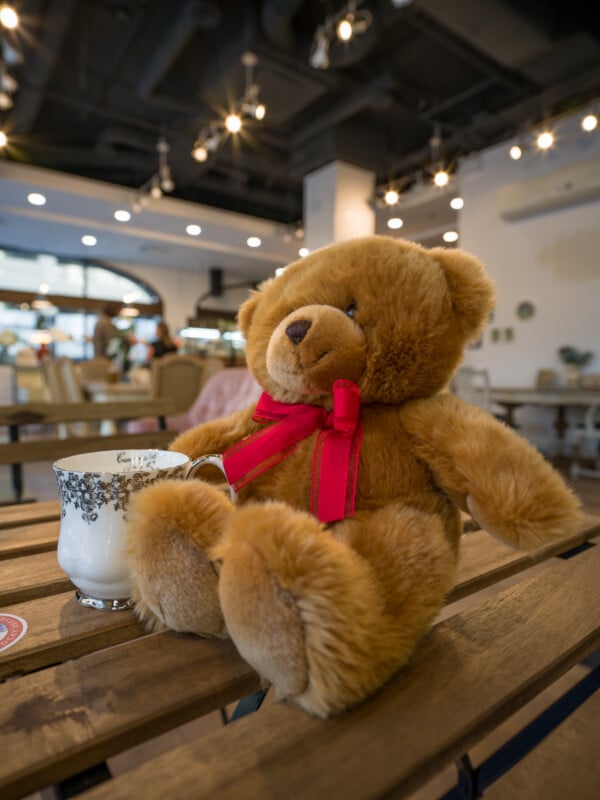 A plush teddy bear with a red bow sitting at a wooden table in a cafe, next to a glass of milk and a decorative cup. the cafe has a modern interior with hanging lights and patrons in the background.