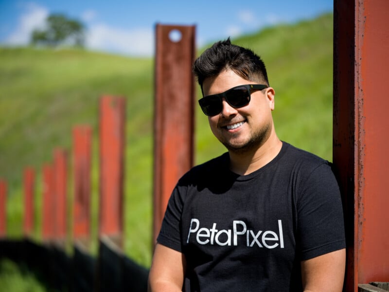 A smiling man wearing sunglasses and a black t-shirt with the logo "petapixel" stands by a red fence with a lush green hill in the background.