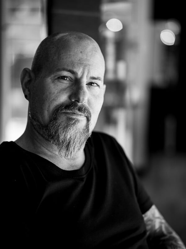 A black and white portrait of a bald man with a beard, looking directly at the camera with a subtle smile, blurred lights in the background.