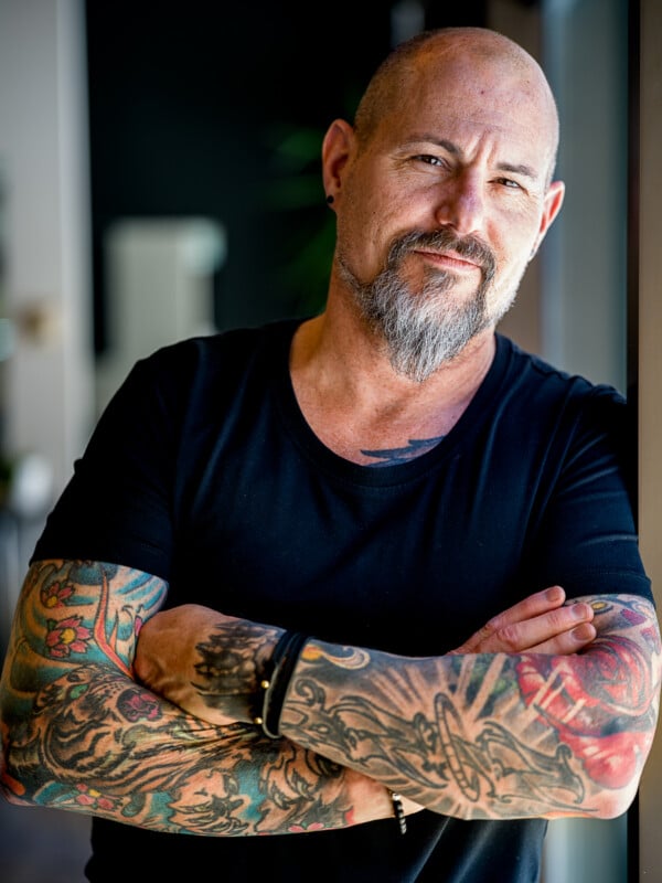 A confident man with a shaved head and a full beard, wearing a black t-shirt, stands with his tattoo-covered arms crossed. he smiles slightly, looking directly at the camera.