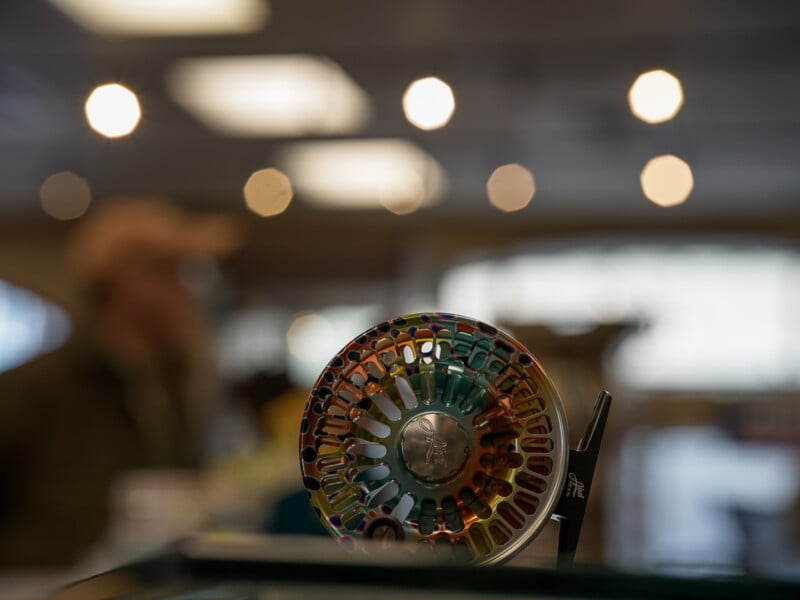 Close-up of a colorful fishing reel on display with soft-focus background showing lights and a blurred person in a hat indoors.