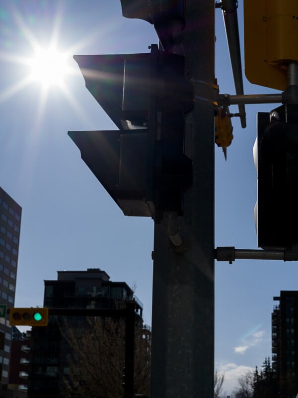 A sunlit image depicting traffic lights and a pedestrian signal on a pole, with a bright sunburst in the clear sky and urban buildings in the background.