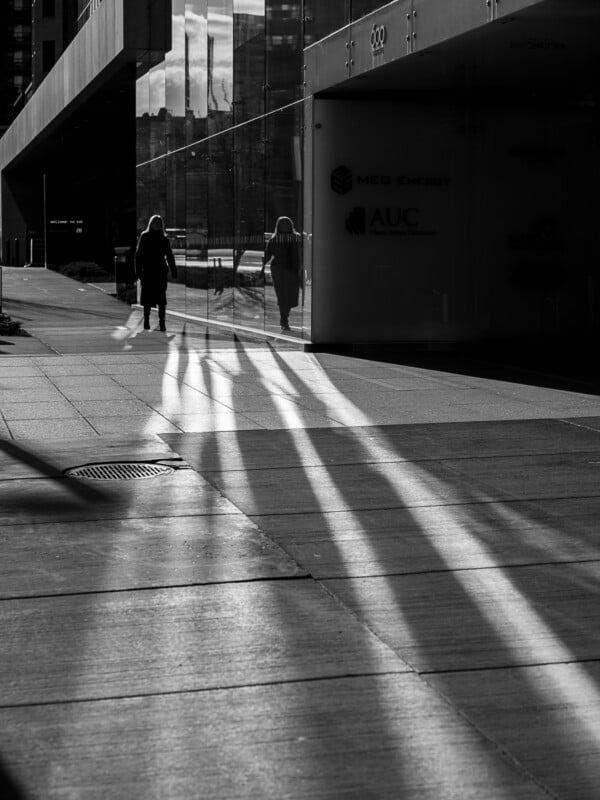 Long shadows stretch across a city sidewalk in black and white, with a person walking away and reflections in the glass building to the right.