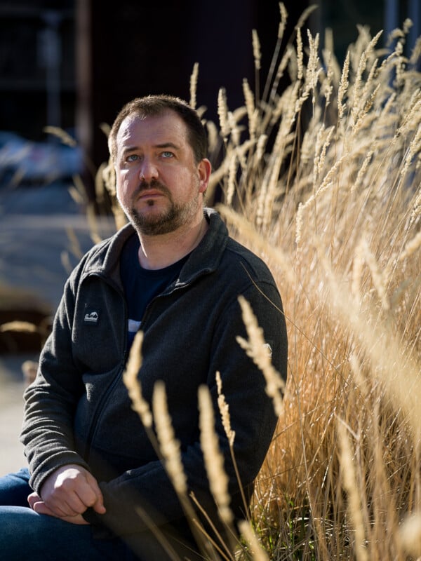 A man with a beard, wearing a dark jacket, sits in front of tall dried grass, looking thoughtfully at the camera, with sunlight filtering through the foliage.