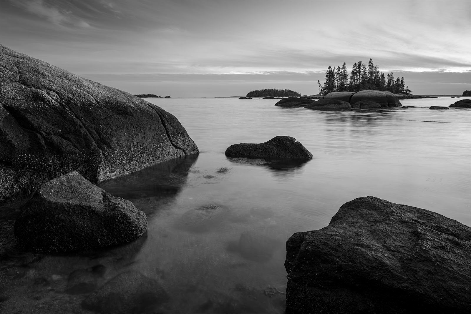 A black and white photo of a serene lake with smooth water, featuring large rocks in the foreground and a small, tree-covered island in the background under a cloudy sky.