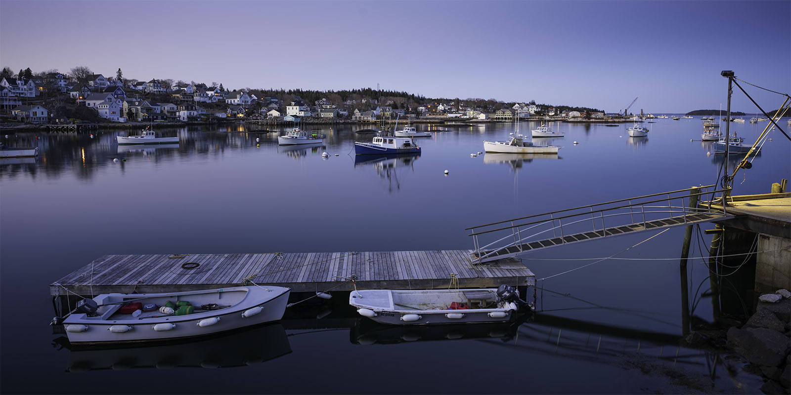 Twilight settles over a serene harbor with boats moored peacefully on calm waters. a small floating dock in the foreground holds two boats, against a backdrop of a quaint hillside town.