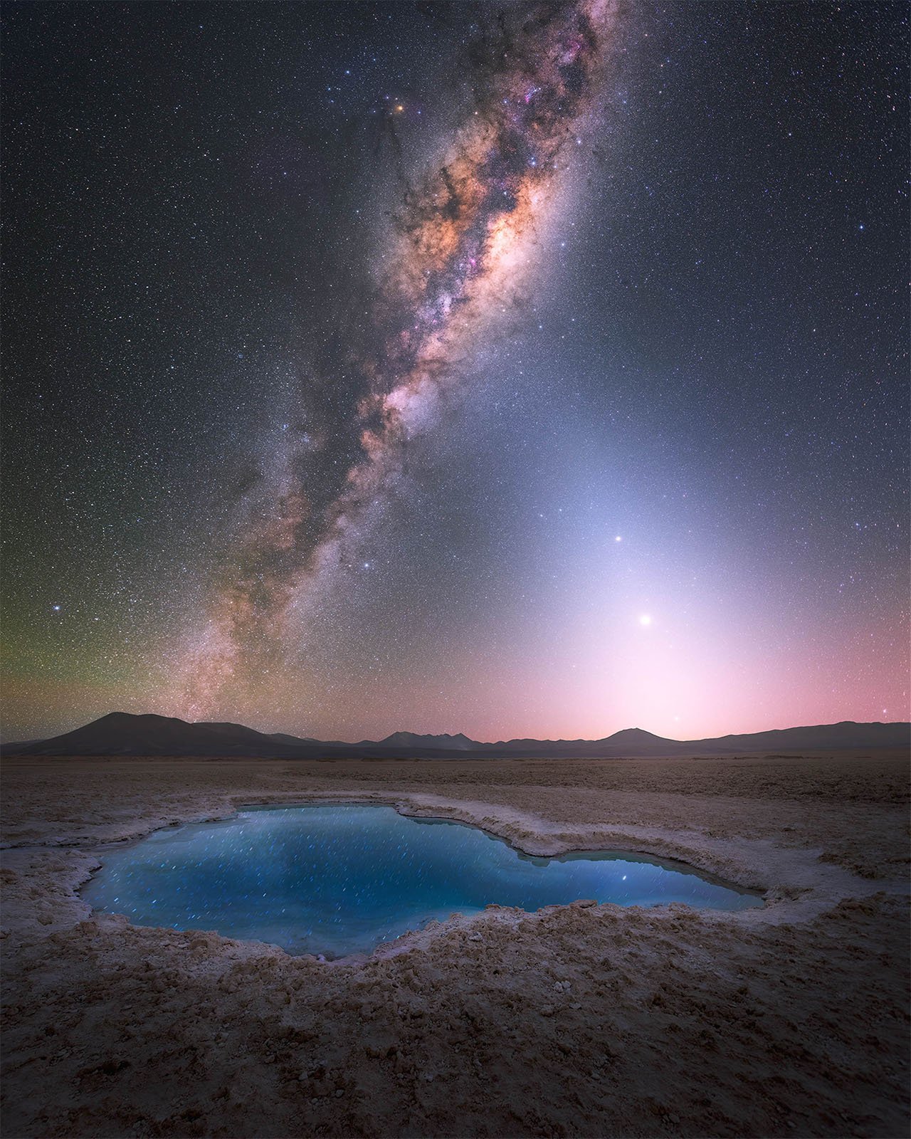  A mesmerizing night sky filled with stars and the Milky Way galaxy stretches above a tranquil desert landscape. Central to the image, a pristine blue pool of water on the ground reflects the starry sky above. Distant mountains frame the horizon under the celestial display.