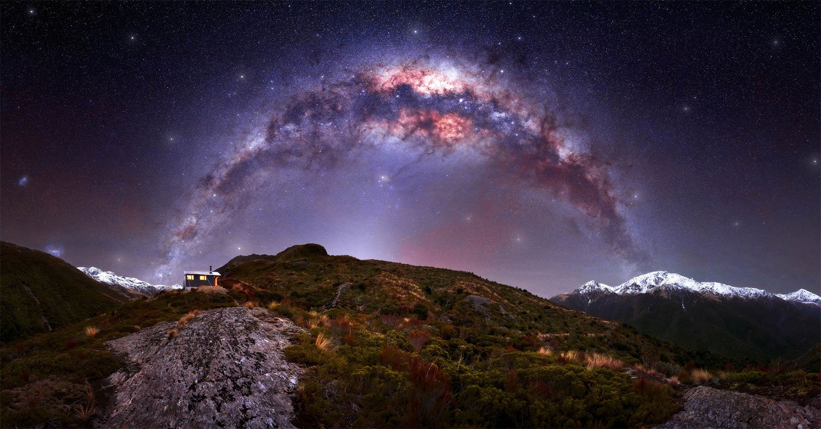 A panoramic view of a starry night sky with the Milky Way arching over rugged, snow-capped mountains. A solitary house with a light on is nestled on a hill in the foreground, surrounded by shrubbery and rocky terrain. The scene conveys tranquility and natural beauty.