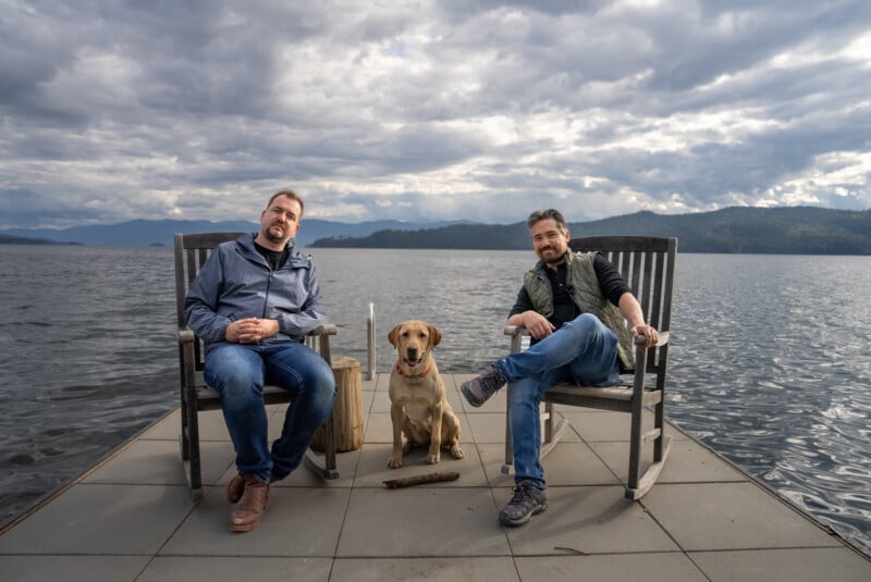 Two men and a golden retriever sitting on a dock by a lake with distant mountains under cloudy skies. one man holds a stick, and the dog looks attentively at the camera.