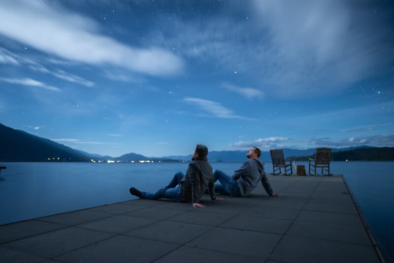 Two people sitting back-to-back on a wooden dock at night, gazing at a starry sky with streaks of clouds over a tranquil lake and distant mountains.