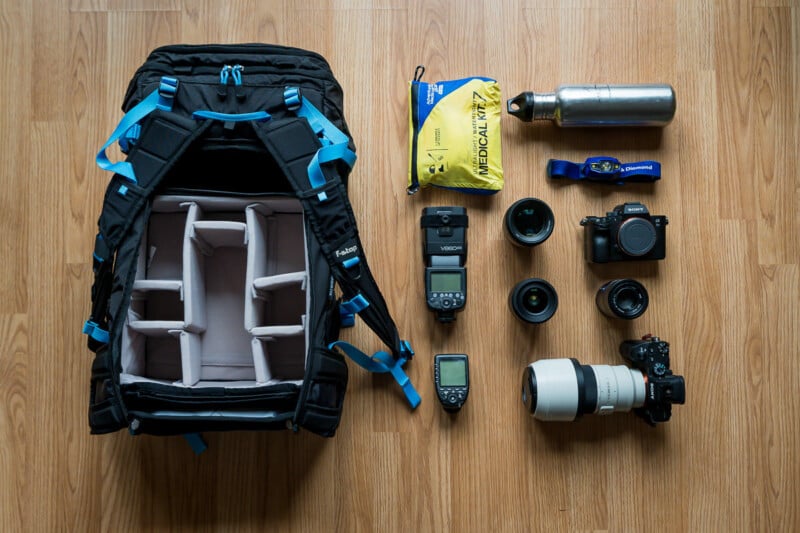 Top view of photography equipment and hiking gear laid out on a wooden floor, including a backpack, camera, lenses, GPS, water bottle, and hydration pack.
