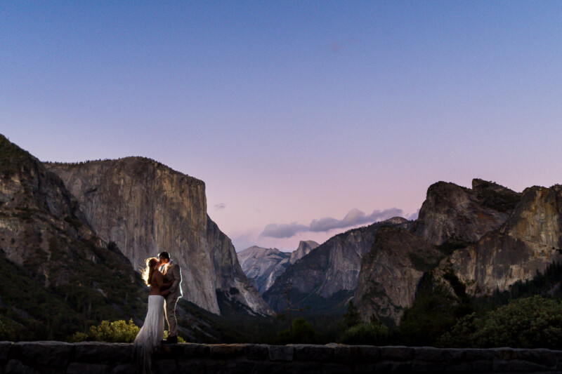 A couple embraces at dusk in Yosemite valley, with the iconic granite cliffs and a soft pink sunset in the background.