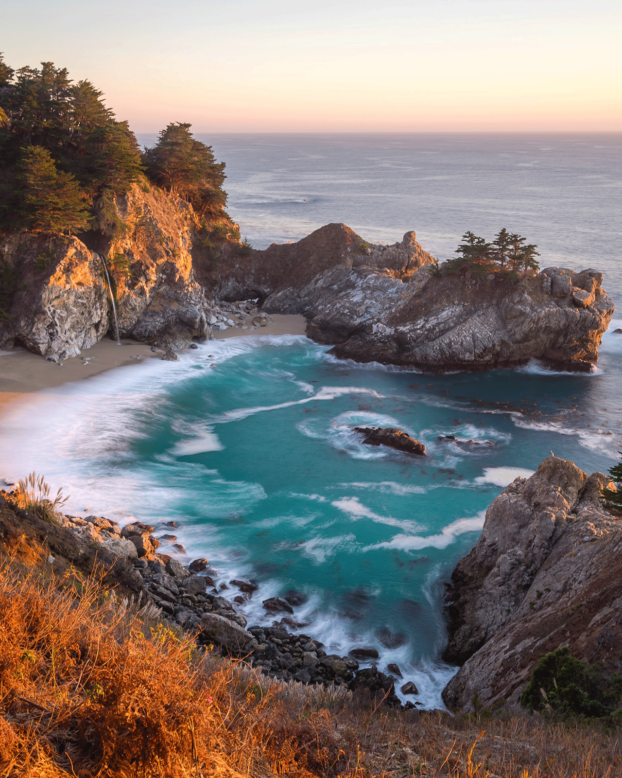 A breathtaking view of a serene, turquoise cove along a rugged coastline at sunset. A gentle waterfall cascades onto the sandy beach, surrounded by rocky cliffs and lush greenery. The sky is painted with warm hues of orange and pink, reflecting on the tranquil water.