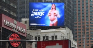 A digital billboard in a cityscape features an advertisement with a pregnant woman holding two bagels over her chest. The text reads, "Just Add Milk with Molly Baz" and includes a website URL "sweetmilk.com/mollybaz." Surrounding the billboard are buildings and a Crown Plaza hotel.
