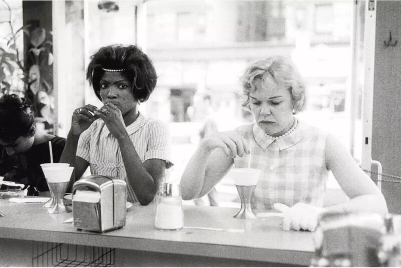 Black and white photo of two women sitting at a diner counter. Both are drinking milkshakes, one with a straw and the other with a spoon. The woman on the right appears to be stirring her shake. Various diner items are on the counter, like a napkin holder and a sugar dispenser.