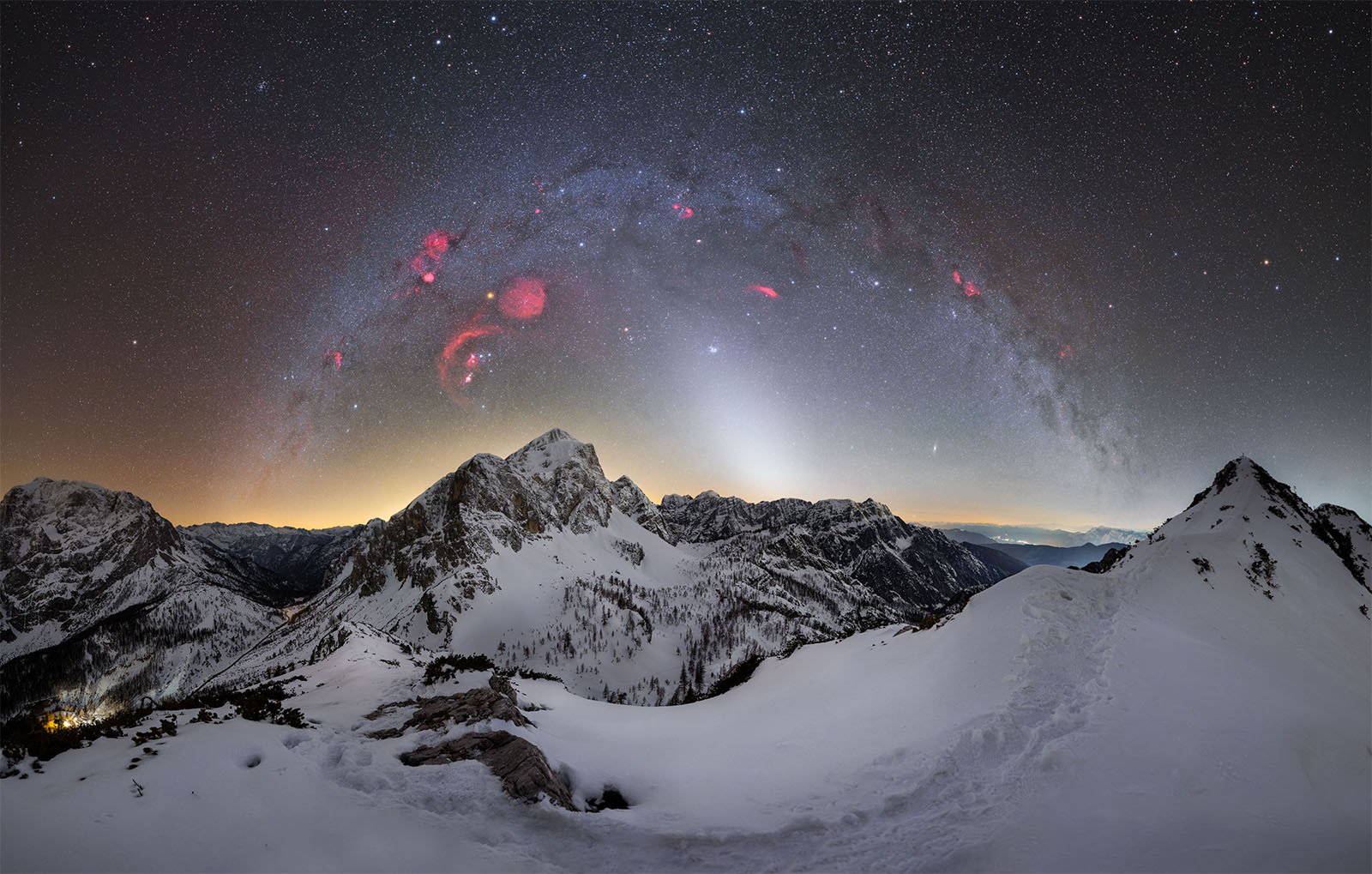 A breathtaking night sky above snow-covered mountains. The Milky Way arcs overhead, with clusters of red nebulae visible. Distant mountain peaks are illuminated by the soft glow of city lights on the horizon, creating a serene and otherworldly landscape.