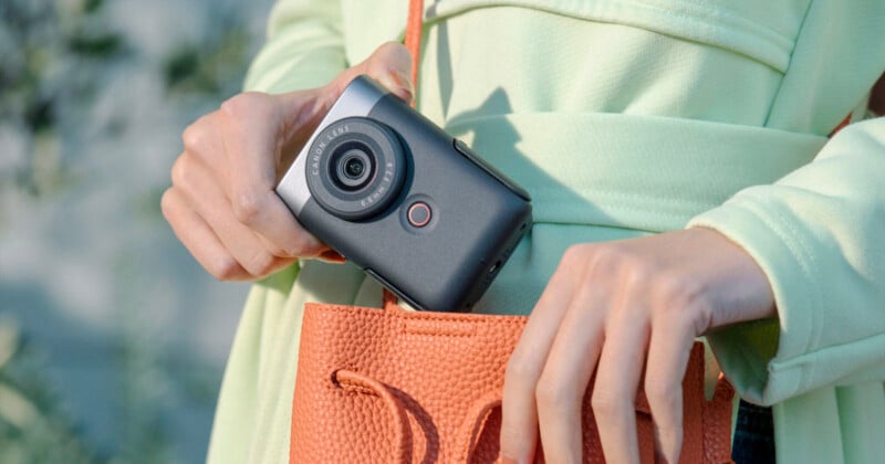 A person is placing a small digital camera into an orange textured purse. The person is wearing a light green shirt and the background is blurred greenery.