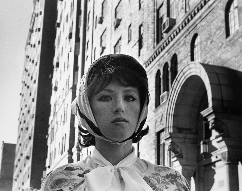 Black and white image of a person standing in front of a tall brick building. They are wearing a scarf tied under their chin and a helmet-like accessory. The building features arched windows and an ornate entrance.