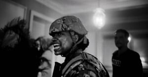 Black and white image of a soldier in camouflage gear and helmet, holding a rifle, shouting or yelling. The background is slightly blurred, featuring another person, a bright hanging light, and some foliage.