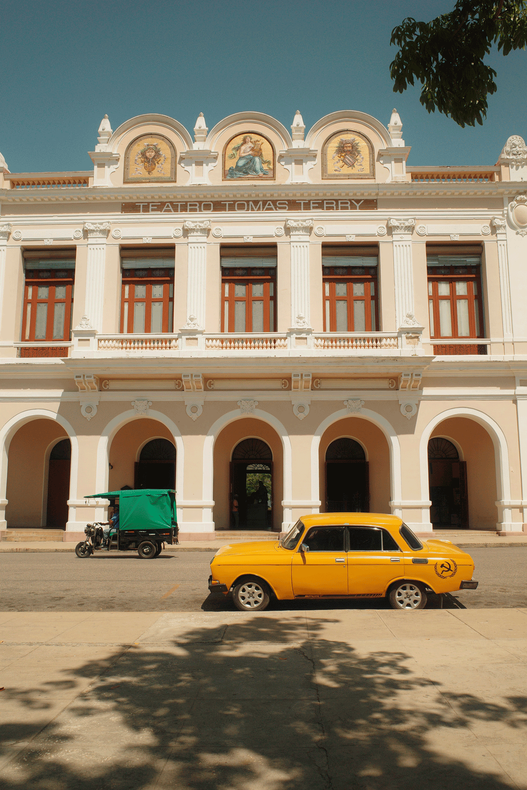 A yellow vintage car with a hammer and sickle symbol is parked in front of Teatro Tomás Terry. The building features three arches, grand columns, and ornate decorations, with three round paintings above. A green and black tuk-tuk is also parked nearby.