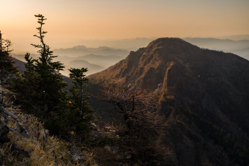A scenic view of a mountain range at sunrise, with a prominent rocky peak in the foreground. Trees with sparse foliage are visible on the slope, and misty layers of distant mountains fade into the horizon, creating a serene and atmospheric landscape.
