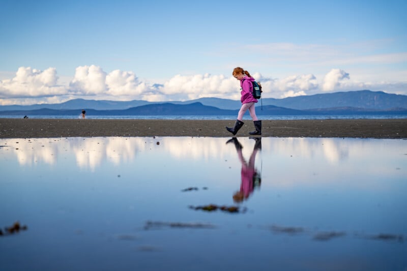 A woman walks along a reflective wet beach, her figure mirrored in the water, with distant mountains and a cloudy sky in the background.