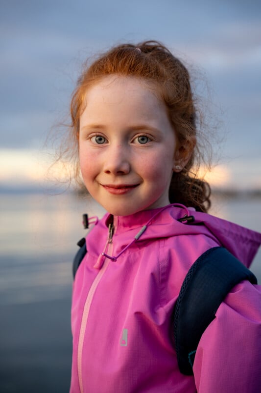 Close-up portrait of a young girl with red hair and freckles, wearing a pink jacket and backpack, smiling gently against a soft-focus background of a lake during twilight.