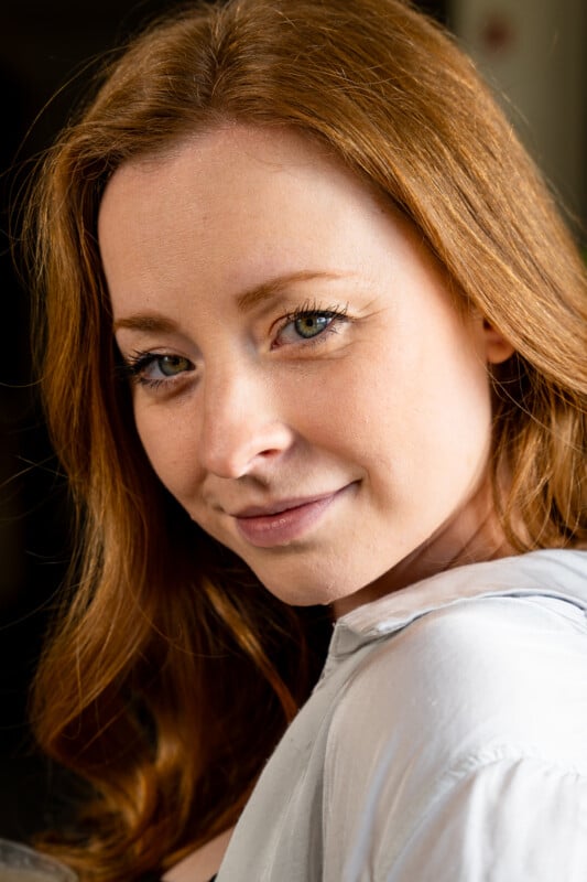 Close-up portrait of a woman with long red hair and subtle makeup, giving a gentle smile to the camera. She wears a white blouse and is softly lit from the side.