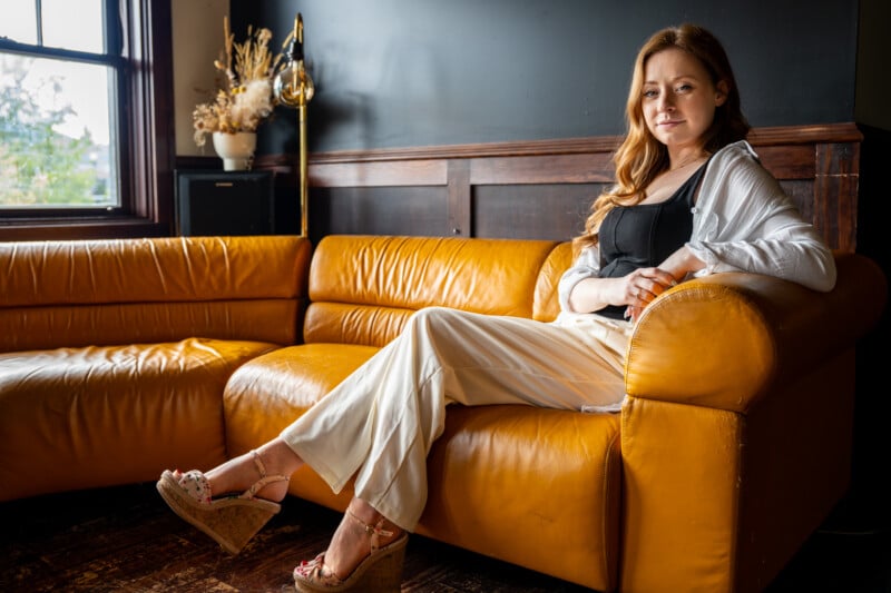 A woman lounging on a mustard yellow leather couch in a stylish room, holding a book and looking thoughtfully at the camera. She wears a white blouse, beige pants, and heeled sandals.