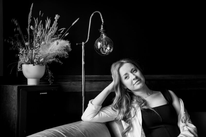 Black and white photo of a woman sitting on a bed, leaning on her hand, with a hanging lightbulb and a vase of dried flowers beside her. She has a contemplative expression.