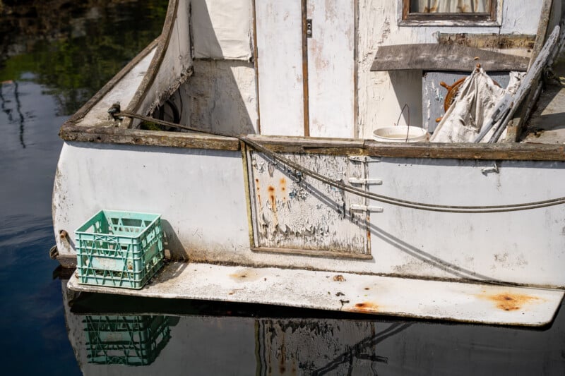 Side view of a worn white boat's stern with a green fishing crate on it, moored to a dock, reflecting clear details in calm water.