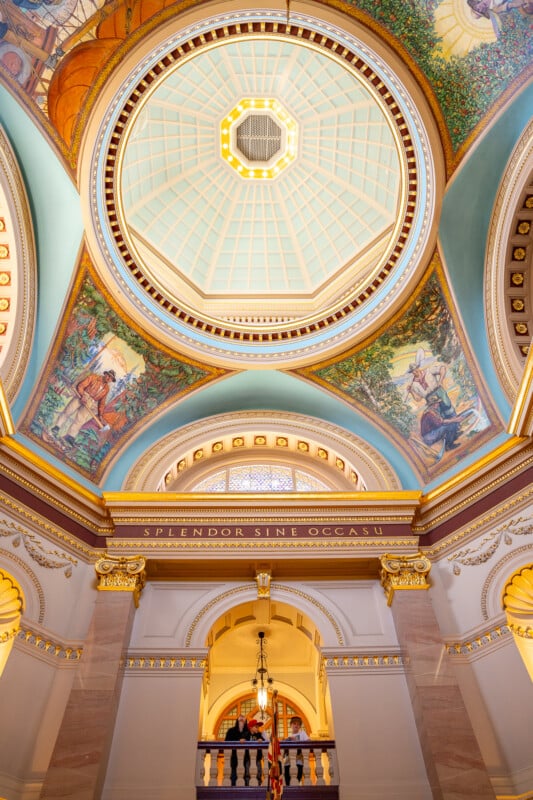 Interior of a grand building featuring a high dome ceiling with a central skylight, ornate frescoes, and classical architectural details. The inscription "SPLENDOR SINE OCCASU" is visible. People are seen on the lower floor.