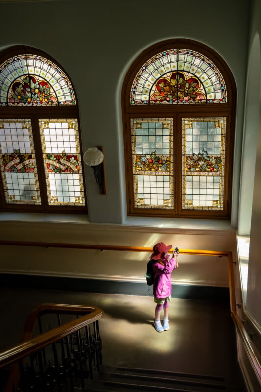 A woman in a pink jacket stands on a stairway, photographing large ornate stained glass windows that are illuminated with natural light.