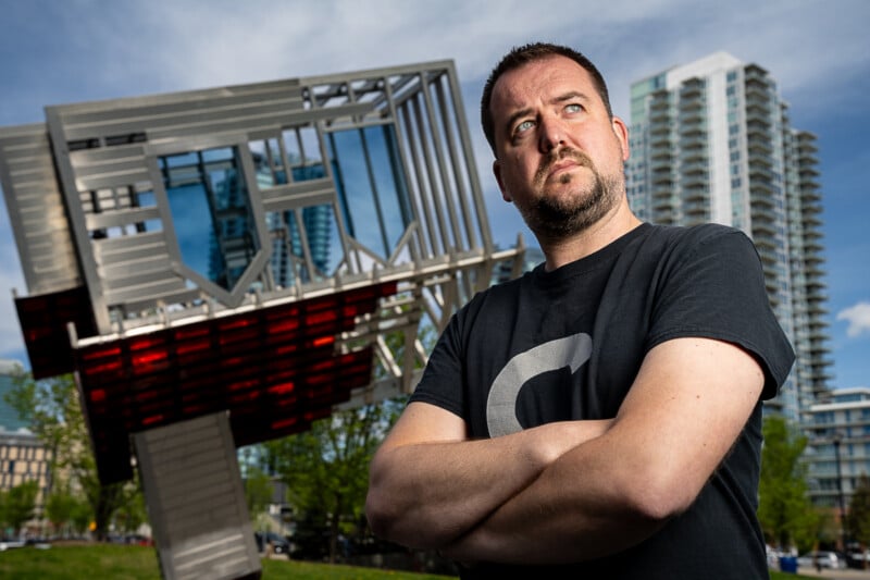 A man with a beard and short hair stands with arms crossed, gazing thoughtfully into the distance. He's wearing a black t-shirt with a large "C" printed on it. Behind him is a modern sculpture and tall buildings against a blue sky with clouds.