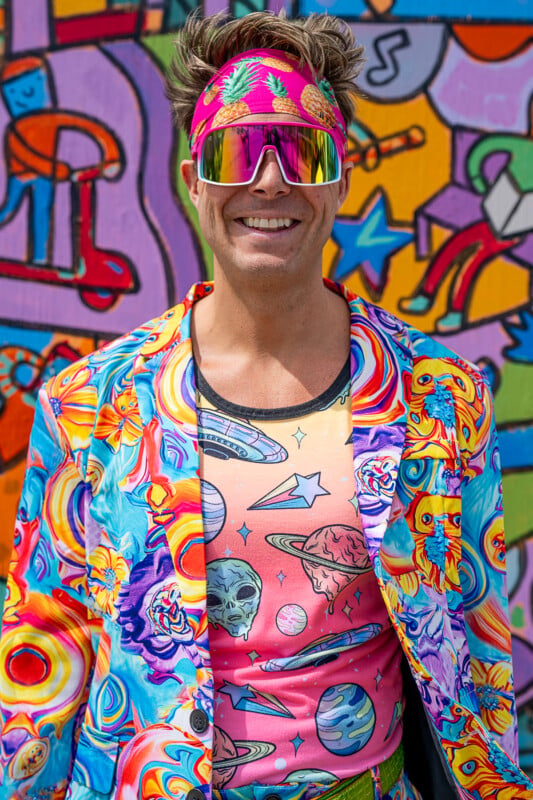 A man is wearing a vibrant, multicolor outfit with swirling, psychedelic patterns, a headband adorned with pineapples, and large reflective sunglasses. The background features a colorful mural with various abstract and cartoonish designs.