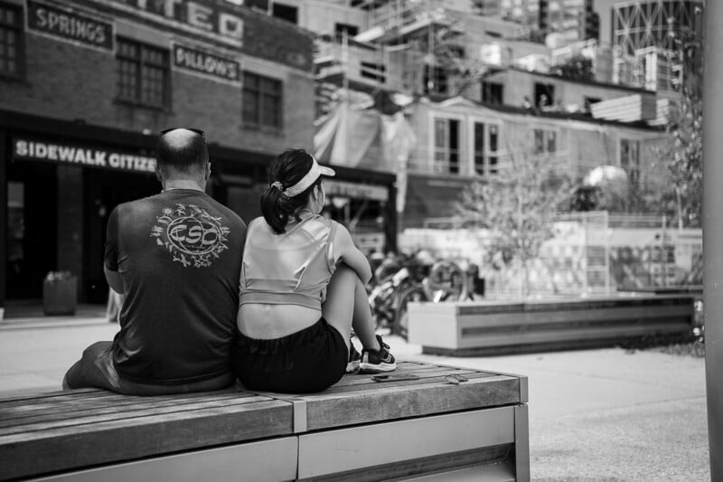 A man and a woman sit side by side on a bench in an urban setting. The man, wearing a T-shirt, and the woman, in a sports outfit and visor, both face away from the camera. Behind them are buildings with visible signage, and a construction area is also in view.